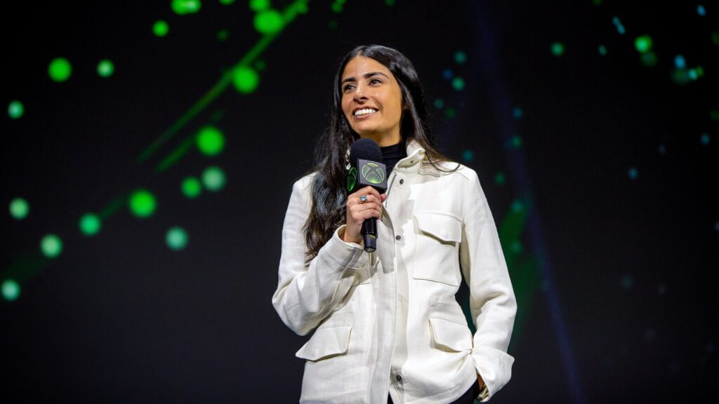 Xbox CEO Asha Sharma Details Plan to Turn Business Around Asha Sharma speaking on stage at an Xbox event with an Xbox-branded microphone and green backdrop