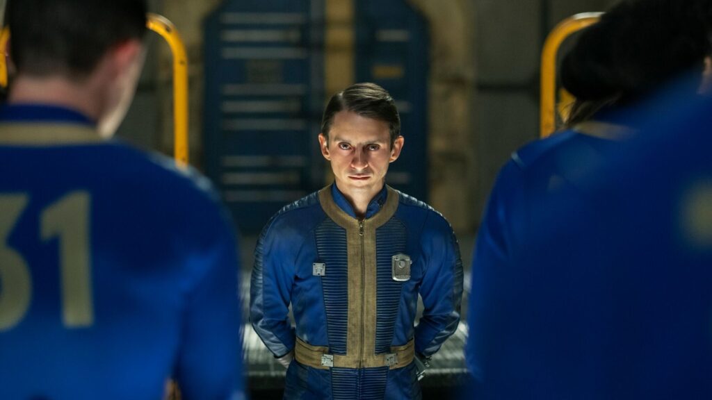 Fallout Shelter Reality Show Premieres January 13 on Prime Video Official Fallout Shelter promotional image showing a man in a blue-and-gold Vault-Tec jumpsuit standing inside a dimly