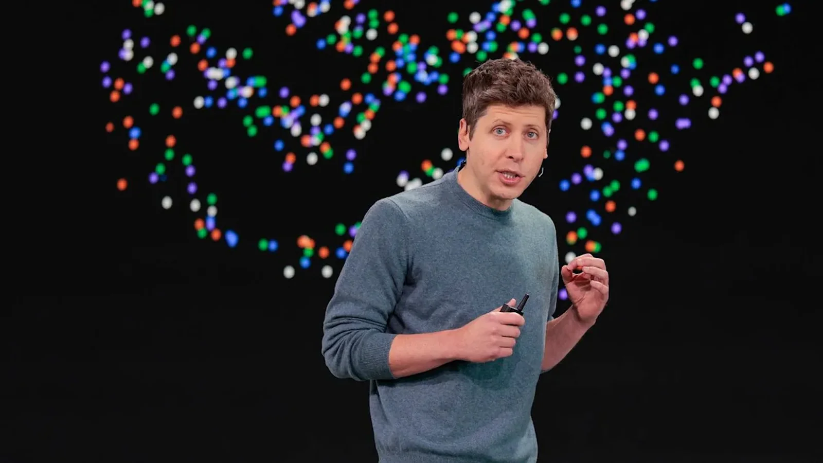 Sam Altman speaking onstage at OpenAI DevDay 2026, wearing a blue-gray sweater against a black background with colorful